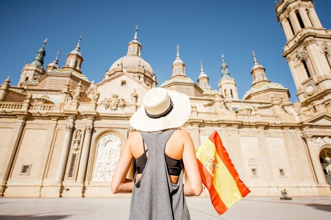 Young woman tourist standing back with spanish flag in front of the famous cathedral on the central square during the sunny weather in Zaragoza city, Spain
