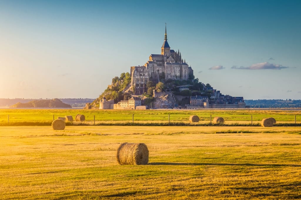 Beautiful view of famous historic Le Mont Saint-Michel tidal island in beautiful golden evening light at sunset in summer with hay bales on empty fields, Normandy, northern France