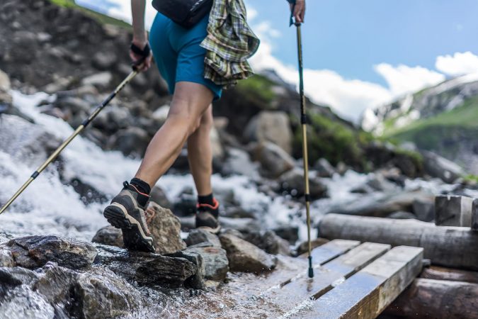 Woman wearing outdoor boots and shorts walking across a wooden bench and rocks crossing a wild alpine creek using hiking poles. Trekking shoes on rocks and in runnig water.