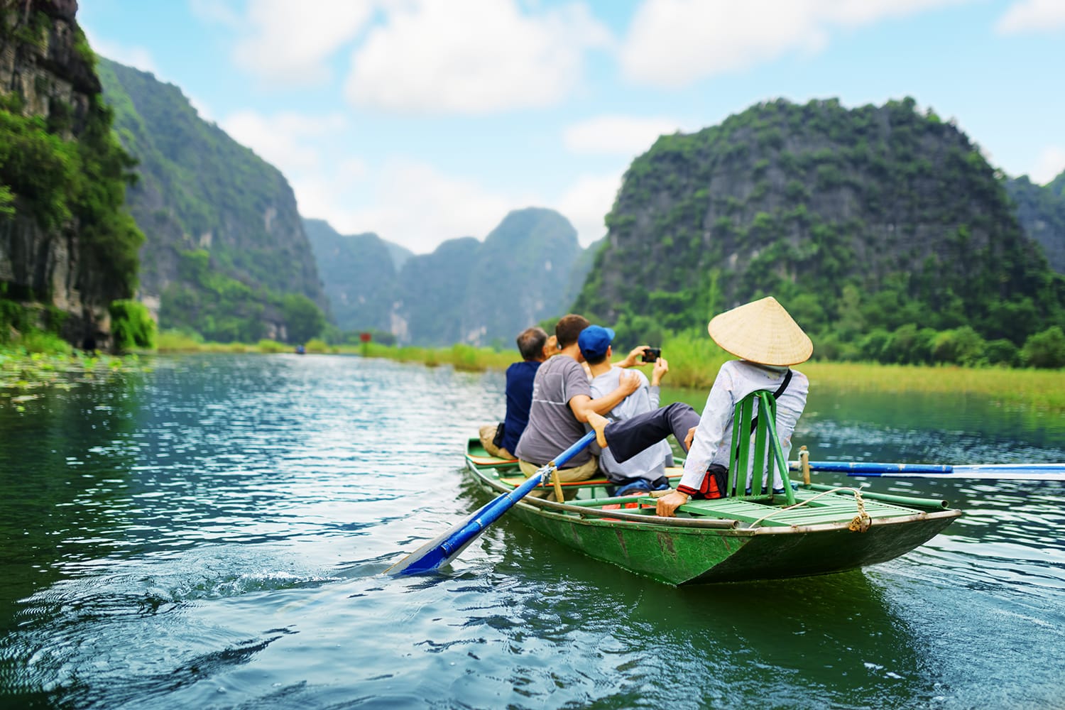 Tourists traveling in boat along the Ngo Dong River and taking picture of the Tam Coc, Ninh Binh, Vietnam. Rower using her feet to propel oars. Landscape formed by karst towers and rice fields.
