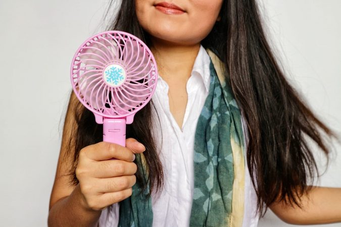 Woman with long brunette hair, wears white shirt with scarf, holding portable pink fan