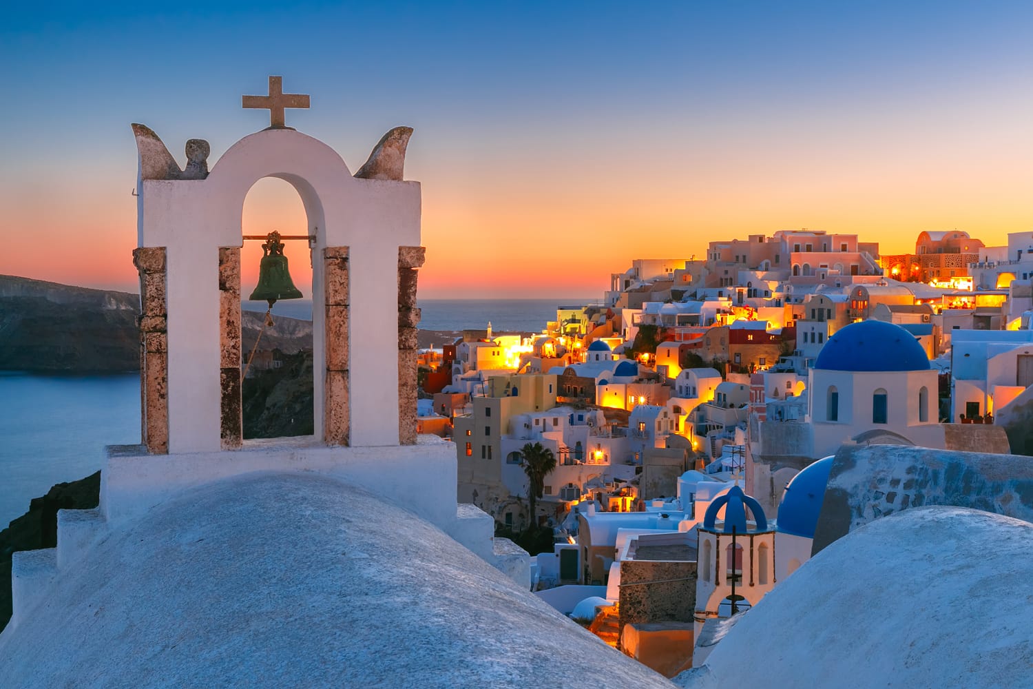 Lengkungan dengan lonceng, rumah putih dan gereja dengan kubah biru di Oia atau Ia saat matahari terbenam keemasan, pulau Santorini, Yunani.
