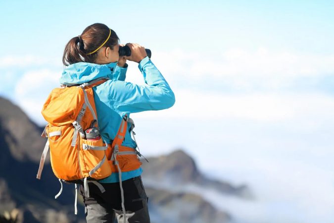 Hiker looking in binoculars enjoying spectacular view on mountain top above the clouds.