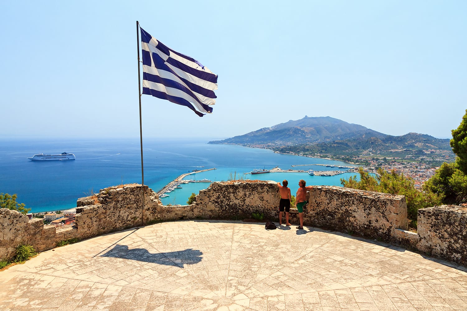 Father and son looking at the city of Zakynthos from the bochali venetian castle in summer with a greek flag