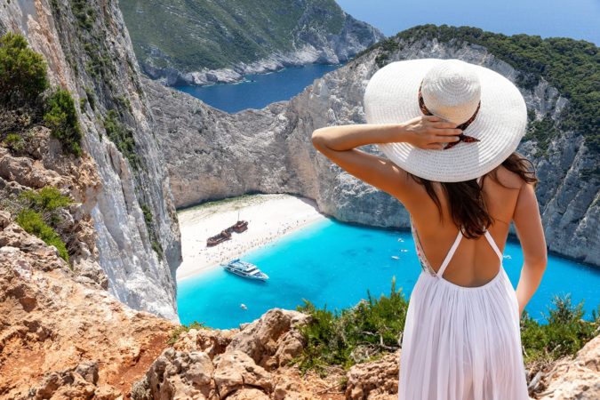 Attractive traveler woman stands on a cliff and enjoys the breathtaking view to the famous shipwreck beach, Navagio, on Zakynthos island, Greece