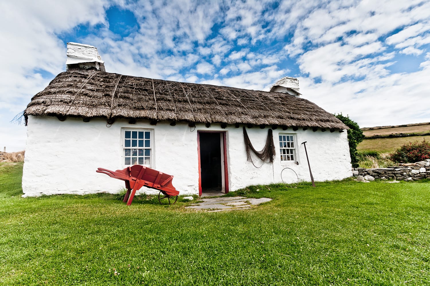 Old fashioned thatched crofter's cottage at Cregneash in the Isle of Man with red wooden wheelbarrow, fishing net and agricultural tool in the foreground with green grass and a blue sky.