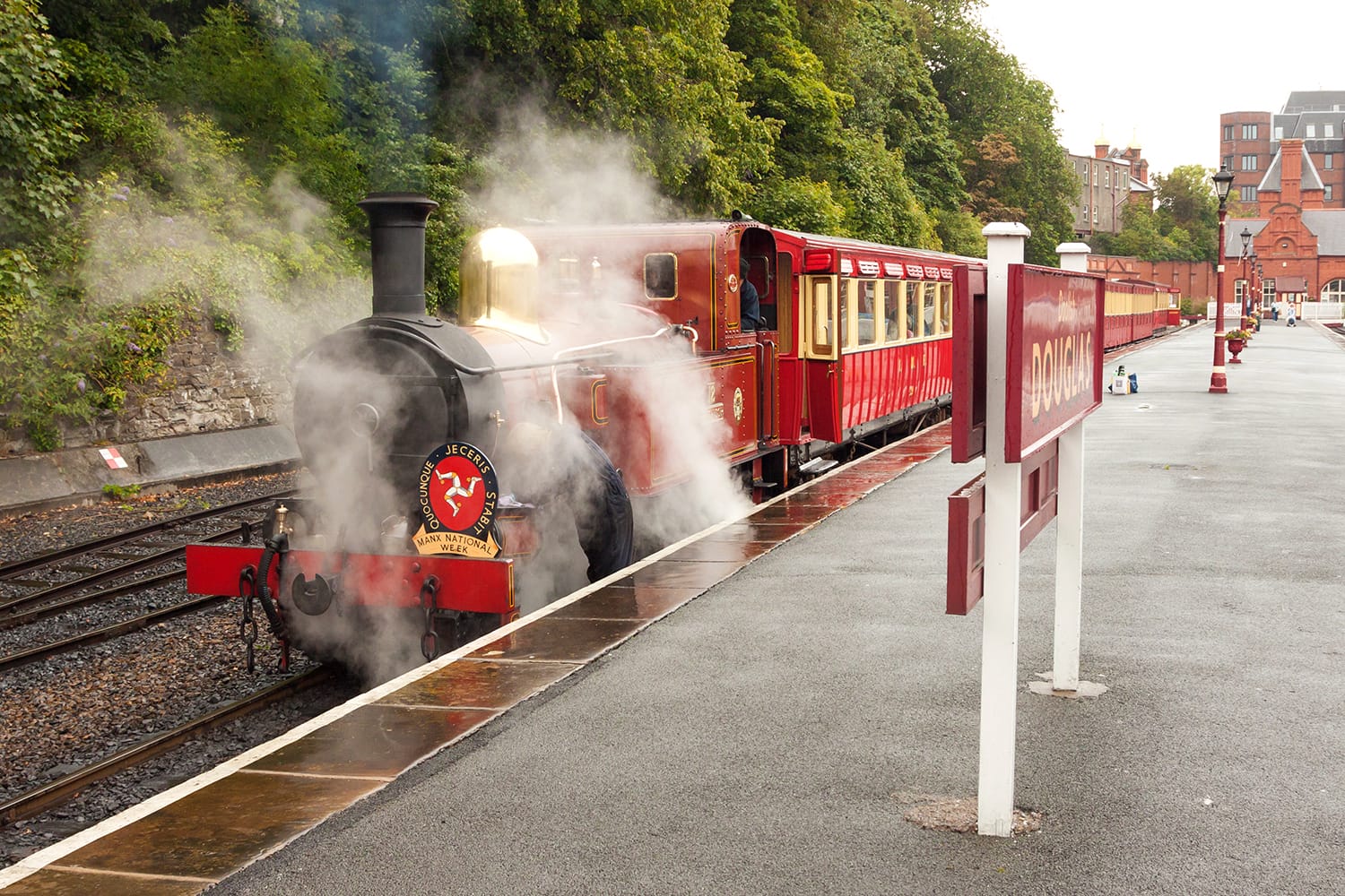 The red steam locomotive with train of IMR line in puffs of vapour prepares to depart from the platform of Douglas railway terminal station