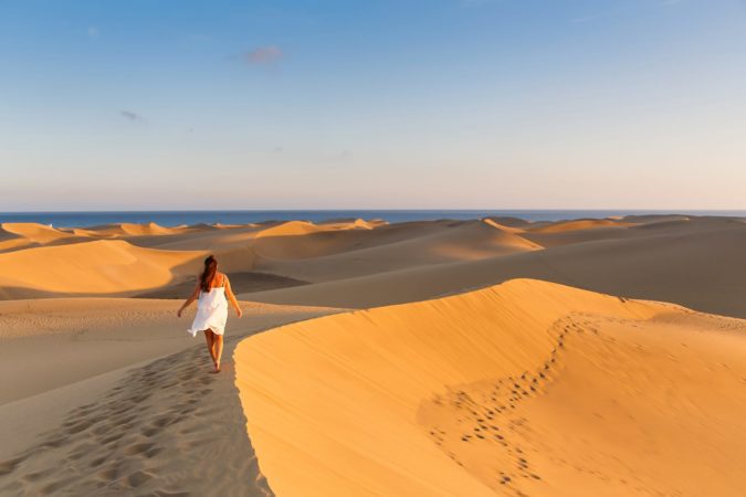 Young beautiful woman walking on the sand wearing white dress at Maspalomas dunes in Gran Canaria, Spain