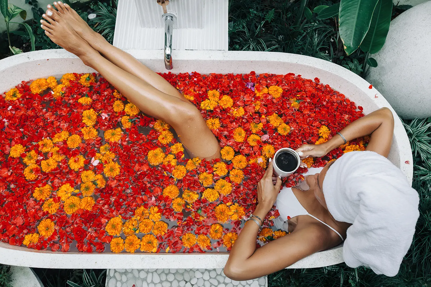 Woman with wrapped towel on head is drinking tea or coffee while relaxing in outdoor bath with flowers in Bali spa hotel, top view from above