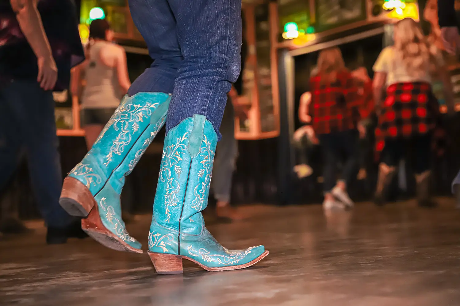 A female wearing turqoise cowboy boots dances at a bar`s country-themed night.