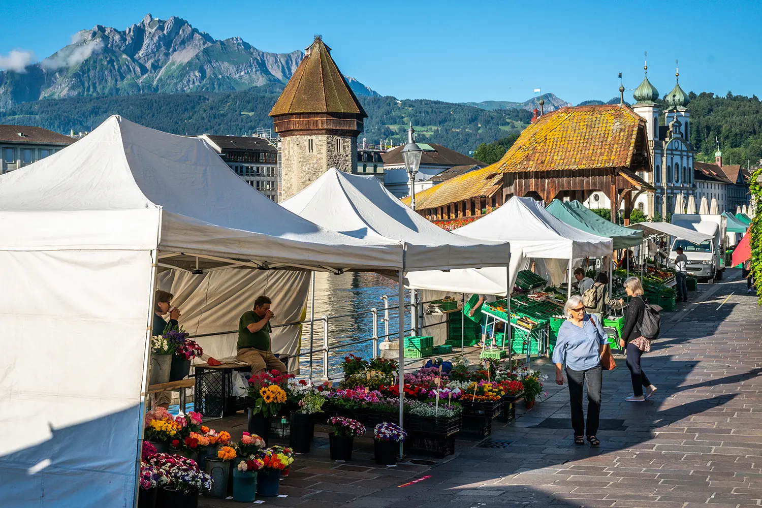 Lucerne morning farmer market with people and stalls with Chapel bridge landmark and Pilatus mount in background during summer in Lucerne Switzerland